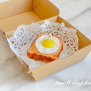 K&ouml;nnte beinhalten: Ein kleines, wei&szlig;es Papierdeckchen liegt in einer braunen Pappbox. Auf dem Deckchen liegt ein kleines, quadratisches St&uuml;ck Brot mit einem Spiegelei darauf. Das Ei ist gelb und das Brot ist braun. Der Text "Smell and Smile" ist auf der Unterseite der Box aufgedruckt.