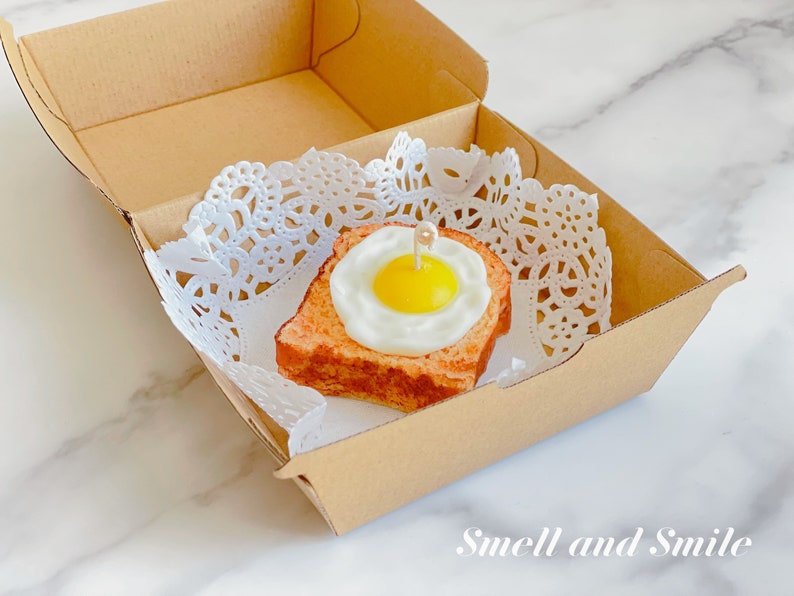 K&ouml;nnte beinhalten: Ein kleines, wei&szlig;es Papierdeckchen liegt in einer braunen Pappbox. Auf dem Deckchen liegt ein kleines, quadratisches St&uuml;ck Brot mit einem Spiegelei darauf. Das Ei ist gelb und das Brot ist braun. Der Text "Smell and Smile" ist auf der Unterseite der Box aufgedruckt.