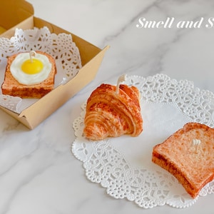 K&ouml;nnte beinhalten: Drei dekorative Kerzen in Form von Fr&uuml;hst&uuml;cksgerichten: ein Croissant, ein Toastbrot und ein Toastbrot mit einem Spiegelei darauf. Die Kerzen stehen auf wei&szlig;en Doilies und der Text "Smell and Smile" ist in Wei&szlig; auf hellbraunem Hintergrund geschrieben.