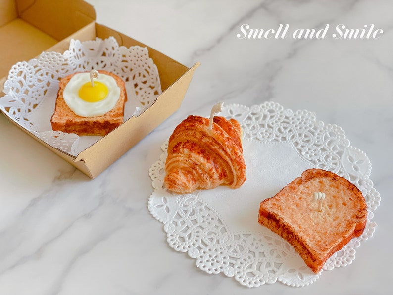 K&ouml;nnte beinhalten: Drei dekorative Kerzen in Form von Fr&uuml;hst&uuml;cksgerichten: ein Croissant, ein Toastbrot und ein Toastbrot mit einem Spiegelei darauf. Die Kerzen stehen auf wei&szlig;en Doilies und der Text "Smell and Smile" ist in Wei&szlig; auf hellbraunem Hintergrund geschrieben.