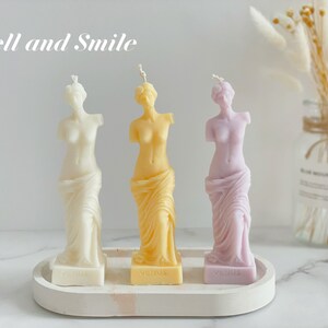 May include: Three decorative candles shaped like the Venus de Milo statue. The candles are white, yellow, and purple. The candles are on a white tray with the word "Venus" printed on each candle base. The text "Smell and Smile" is printed above the candles.