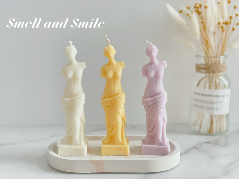 May include: Three decorative candles shaped like the Venus de Milo statue. The candles are white, yellow, and purple. The candles are on a white tray with the word "Venus" printed on each candle base. The text "Smell and Smile" is printed above the candles.