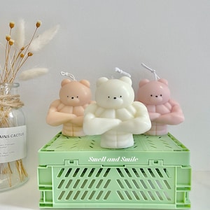 Peut inclure: Trois bougies parfumées en forme d'ours musclés. Les ours sont en rose, blanc et marron. Les bougies sont posées sur une caisse verte avec les mots "Smell and Smile" imprimés sur le devant.