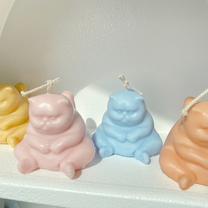 May include: Four hand-poured candles shaped like cats in yellow, pink, blue, and orange. Each candle has a cotton wick and is sitting on a white surface.