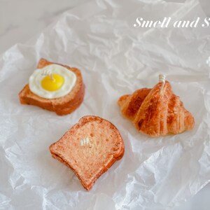 K&ouml;nnte beinhalten: Drei Lebensmittel-f&ouml;rmige Kerzen auf einem wei&szlig;en Papierhintergrund. Die Kerzen sind in Form eines Croissants, einer Scheibe Toast und einer Scheibe Toast mit einem Spiegelei darauf geformt. Der Text "Smell and Smile" steht in der oberen rechten Ecke des Bildes.