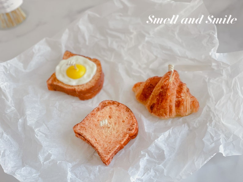 K&ouml;nnte beinhalten: Drei Lebensmittel-f&ouml;rmige Kerzen auf einem wei&szlig;en Papierhintergrund. Die Kerzen sind in Form eines Croissants, einer Scheibe Toast und einer Scheibe Toast mit einem Spiegelei darauf geformt. Der Text "Smell and Smile" steht in der oberen rechten Ecke des Bildes.