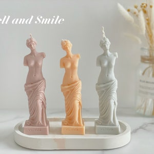 May include: Three decorative candles shaped like the Venus de Milo statue. The candles are in shades of pink, peach, and light blue. Each candle has the word "Venus" printed on the base.