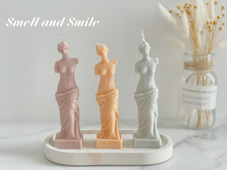 May include: Three decorative candles shaped like the Venus de Milo statue. The candles are in shades of pink, peach, and light blue. Each candle has the word "Venus" printed on the base.