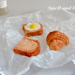 K&ouml;nnte beinhalten: Drei Kerzen in Form von Fr&uuml;hst&uuml;cksgerichten: ein Croissant, eine Scheibe Toast und eine Scheibe Toast mit einem Spiegelei darauf. Die Kerzen stehen auf einem wei&szlig;en St&uuml;ck Pergamentpapier. Der Text "Smell and Smile" ist oben im Bild in Schwarz geschrieben.