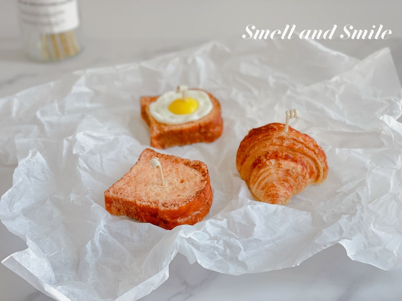K&ouml;nnte beinhalten: Drei Kerzen in Form von Fr&uuml;hst&uuml;cksgerichten: ein Croissant, eine Scheibe Toast und eine Scheibe Toast mit einem Spiegelei darauf. Die Kerzen stehen auf einem wei&szlig;en St&uuml;ck Pergamentpapier. Der Text "Smell and Smile" ist oben im Bild in Schwarz geschrieben.