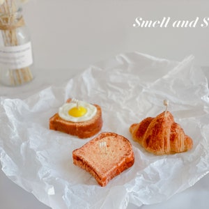 K&ouml;nnte beinhalten: Drei kleine, realistisch aussehende Lebensmittelkerzen auf einem wei&szlig;en Pergamentpapier-Hintergrund. Die Kerzen sind in Form eines Croissants, einer Scheibe Brot mit einem Spiegelei darauf und einer Scheibe Toast geformt. Der Text "Smell and Smile" ist in Wei&szlig; auf einem hellgrauen Hintergrund geschrieben.