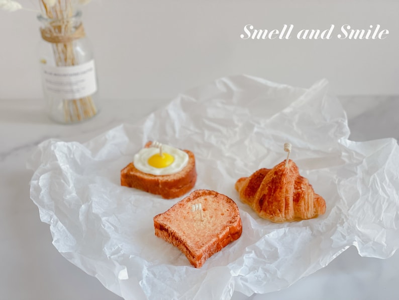K&ouml;nnte beinhalten: Drei kleine, realistisch aussehende Lebensmittelkerzen auf einem wei&szlig;en Pergamentpapier-Hintergrund. Die Kerzen sind in Form eines Croissants, einer Scheibe Brot mit einem Spiegelei darauf und einer Scheibe Toast geformt. Der Text "Smell and Smile" ist in Wei&szlig; auf einem hellgrauen Hintergrund geschrieben.