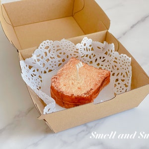 K&ouml;nnte beinhalten: Eine braune und wei&szlig;e Kerze in Form einer Scheibe Brot in einer kleinen Pappbox mit einer wei&szlig;en Spitze. Die Kerze hat einen wei&szlig;en Docht.
