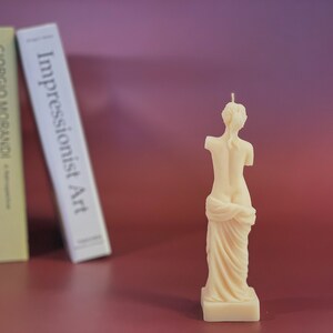 May include: A white candle shaped like a statue of a woman standing with her arms crossed and a draped cloth around her waist.