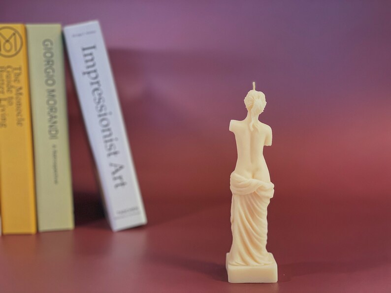May include: A white candle shaped like a statue of a woman standing with her arms crossed and a draped cloth around her waist.