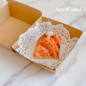 K&ouml;nnte beinhalten: Eine kleine, braune, croissantf&ouml;rmige Kerze in einer Kartonbox mit einer wei&szlig;en Spitze. Die Kerze hat einen wei&szlig;en Docht und ist mit einem braunen &Uuml;berzug verziert. Der Text "Smell and Smile" ist in Wei&szlig; auf der Box geschrieben.