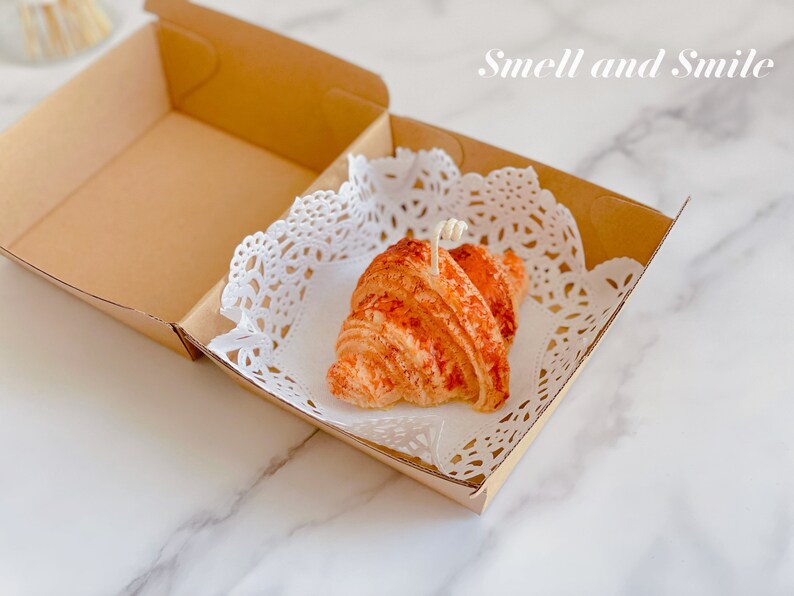 K&ouml;nnte beinhalten: Eine kleine, braune, croissantf&ouml;rmige Kerze in einer Kartonbox mit einer wei&szlig;en Spitze. Die Kerze hat einen wei&szlig;en Docht und ist mit einem braunen &Uuml;berzug verziert. Der Text "Smell and Smile" ist in Wei&szlig; auf der Box geschrieben.