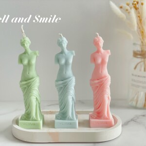 May include: Three decorative candles shaped like the Venus de Milo statue. The candles are green, blue, and pink. The candles are on a white tray with the word "Venus" written on each candle base. The text "Smell and Smile" is written above the candles.