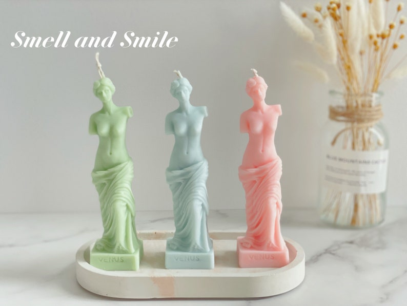 May include: Three decorative candles shaped like the Venus de Milo statue. The candles are green, blue, and pink. The candles are on a white tray with the word "Venus" written on each candle base. The text "Smell and Smile" is written above the candles.