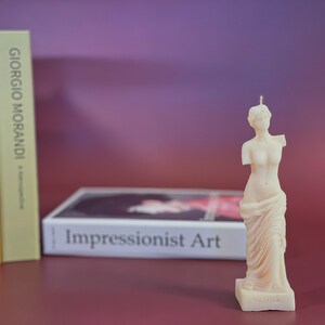 May include: A white candle shaped like the Venus de Milo statue. The candle is on a small base with the word "Venus" inscribed on it. The candle is in front of a book titled "Impressionist Art".