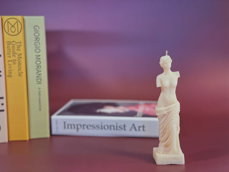 May include: A white candle shaped like the Venus de Milo statue. The candle is on a small base with the word "Venus" inscribed on it. The candle is in front of a book titled "Impressionist Art".
