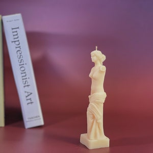 May include: A white candle shaped like a statue of a woman. The candle is on a white base and has a wick on top. The candle is in front of a bookshelf with three books.