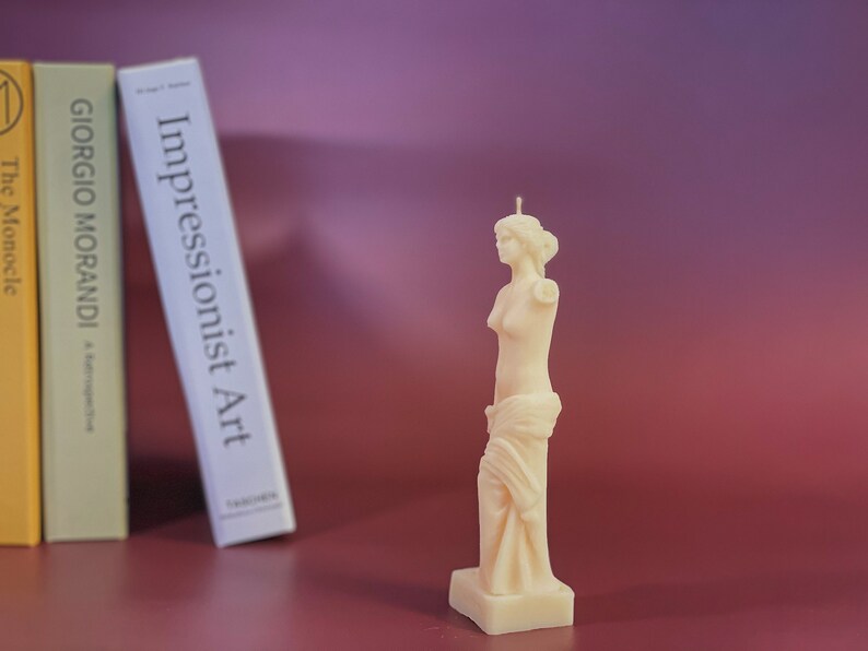 May include: A white candle shaped like a statue of a woman. The candle is on a white base and has a wick on top. The candle is in front of a bookshelf with three books.