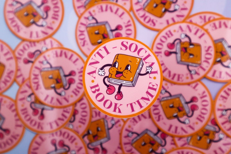 May include: A close-up of several pink and orange round stickers with the text "Anti-Social Book Time" and a cartoon book character with arms and legs.