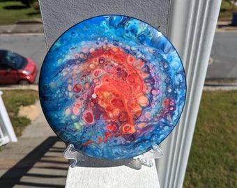 May include: A round disc with a colourful abstract design. The disc is painted with blue, red, orange, yellow, and white colours. The design is reminiscent of a galaxy or nebula. The disc is on a clear plastic stand.