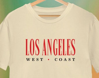 May include: A cream-coloured t-shirt with a red graphic that says "LOS ANGELES" and "WEST COAST" in a vintage style font.