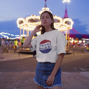 May include: A white t-shirt with a red, white, and blue circle graphic that says "Dead Inside". The person wearing the shirt is standing in front of a carousel at an amusement park.