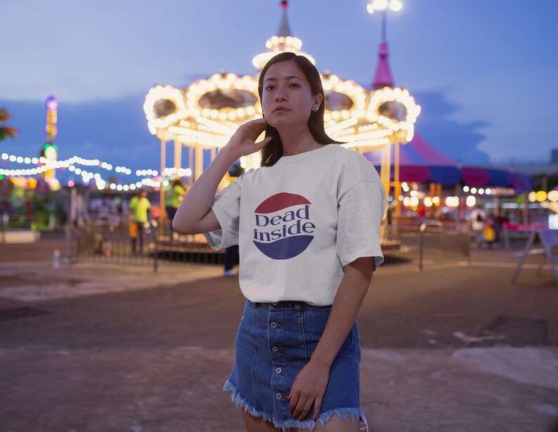 May include: A white t-shirt with a red, white, and blue circle graphic that says "Dead Inside". The person wearing the shirt is standing in front of a carousel at an amusement park.