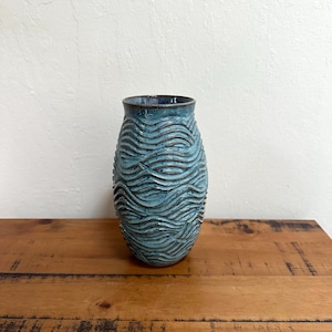 May include: A handcrafted, blue ceramic vase with a textured, wave-like pattern. The vase is cylindrical with a slightly flared top and sits on a wooden surface. The color is a blend of blues, creating a unique decorative piece.