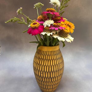 May include: A ceramic vase with a yellow rim and a brown body featuring a repeating vertical oval pattern. The vase is filled with a colorful bouquet of fresh flowers, including pink, white, and orange zinnias and green foliage stems.