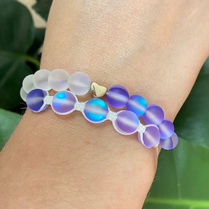 Op de afbeelding: Twee armbanden met kralen. Eén armband heeft heldere, matte kralen. De andere armband heeft iriserende paarse kralen met een gouden hartbedel. Beide armbanden zijn geregen op wit koord.