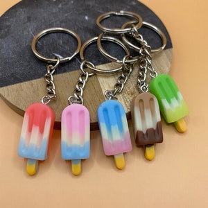 Op de afbeelding: Zes kleurrijke ijslolly sleutelhangers met zilveren sleutelringen. De ijslolly's zijn gemaakt van polymer clay en hebben verschillende kleurcombinaties, waaronder roze, blauw, wit, rood, groen, bruin en geel.