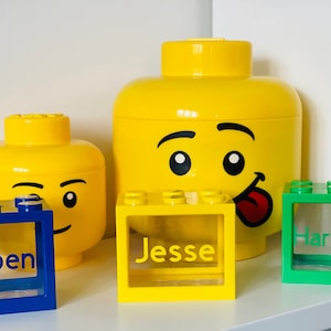 May include: Three yellow Lego head shaped containers with clear plastic windows. The largest container has a smiling face with a tongue sticking out. The other two containers have a winking face. The containers are labelled "Jesse", "Torben", and "Harrison".