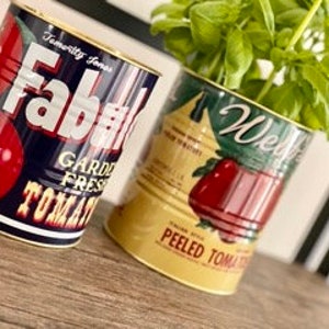 May include: Two vintage-style metal tomato cans repurposed as planters. The cans are yellow, green, and blue with red tomato illustrations and text that reads "Fabrik" and "Weibels".