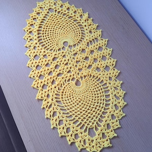 May include: A handmade, yellow crochet doily with a spiral design. The oval-shaped doily features an open, lacy pattern with a scalloped edge. The bright yellow colour adds a touch of warmth and charm to any space.
