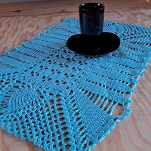 Puede incluir: Un tapete rectangular de ganchillo en color azul claro con un intrincado patrón calado y borde festoneado. Una taza y un platillo de cerámica negra lisa están colocados en el centro de la superficie textil texturizada y cosida a mano.