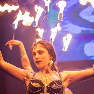 Könnte beinhalten: Eine Frau trägt einen schwarzen BH und passende Höschen mit goldenen Ketten und einer Feuerkrone über dem Kopf. Sie tanzt mit erhobenen Armen und einem Tattoo auf ihrem Oberschenkel.