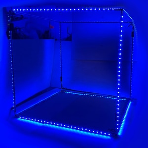 Puede incluir: Una estructura cuadrada de marco abierto iluminada con luces LED azules brillantes. Las luces están dispuestas a lo largo de los bordes del marco, creando un contorno brillante. El interior es oscuro, con un indicio de fondo visible. El efecto general es un diseño moderno e inspirado en la tecnología.