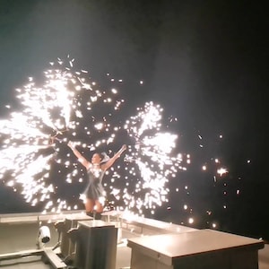May include: A woman in a silver dress stands on a boat deck, holding two sparklers that are creating a shower of sparks.