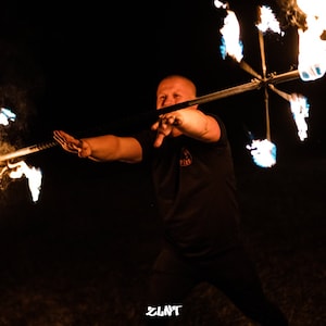 May include: A person performing with a fire staff at night. The staff has multiple flaming elements, creating a dynamic display of light and motion. The background is dark, emphasising the fiery performance.