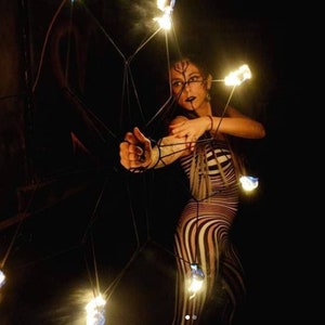 May include: A person in a striped bodysuit and face paint, holding a geometric structure with flames at the ends. The scene is dark, with the flames providing the primary light source, creating a dramatic effect.