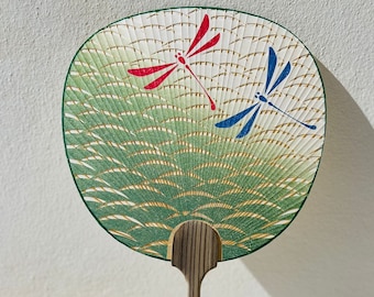 Peut inclure: Un éventail pliant vert et or avec une poignée en bois. L'éventail présente deux libellules, une rouge et une bleue, sur un fond blanc.