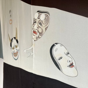 K&ouml;nnte beinhalten: Ein wei&szlig;er Stoff mit drei darauf gemalten japanischen Noh-Masken. Die Masken sind in Schwarz, Wei&szlig; und Rot gehalten. Der Stoff ist Teil eines Kimonos.