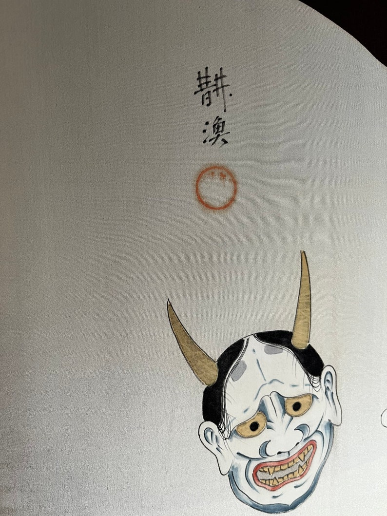 K&ouml;nnte beinhalten: Ein wei&szlig;er Hintergrund mit einer schwarz-wei&szlig;en japanischen D&auml;monenmaske mit goldenen H&ouml;rnern. Die Maske hat ein breites, zahniges Grinsen und gelbe Augen. Der Text "舞" und "楽" ist in schwarzer Tinte &uuml;ber der Maske geschrieben.