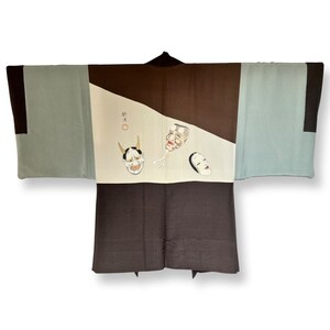 K&ouml;nnte beinhalten: Ein braunes und blaues japanisches Kimono mit einem wei&szlig;en Feld, auf dem drei bemalte Masken abgebildet sind. Die Masken sind eine rote D&auml;monenmaske, eine wei&szlig;e Maske mit einem l&auml;chelnden Gesicht und eine wei&szlig;e Maske mit einem neutralen Ausdruck. Das Kimono hat einen schwarzen Kragen und eine schwarze Sch&auml;rpe.