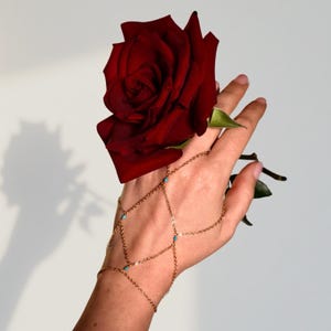 Puede incluir: Una cadena de mano dorada con pequeños detalles turquesas se lleva en una mano que sostiene una gran rosa roja oscura. La cadena es delicada y se conecta a la muñeca, los dedos y el dorso de la mano.